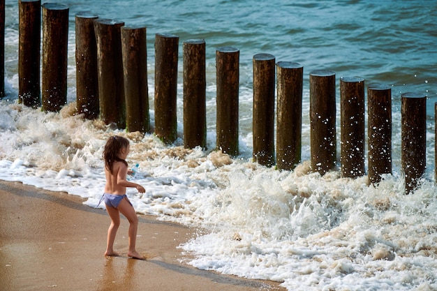 Menina feliz brincando e correndo na praia pelas ondas do mar durante as férias. Criança ativa em calção de banho se divertindo nas ondas do mar no verão. Garota empolgante brincando perto de quebra-mares