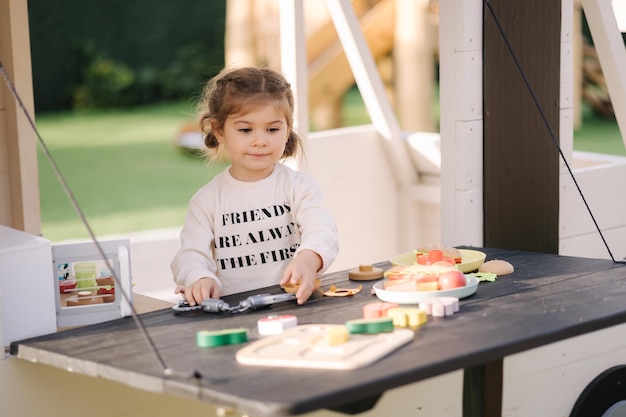 Menina feliz brincando na cozinha de brinquedo sobre rodas linda garota faz um hambúrguer na cozinha de brinquedo ao ar livre