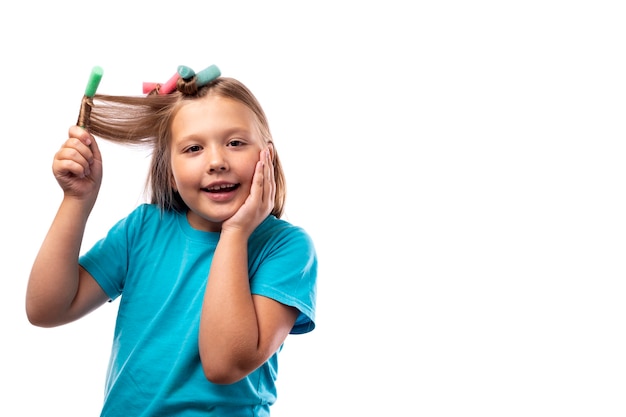 Menina feliz caucasiana em uma camiseta azul enrola seus longos cabelos loiros em bobes sobre um fundo claro. Enrole o cabelo com cachos multicoloridos