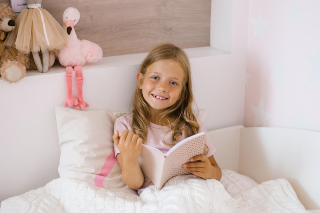 Menina feliz com cabelo comprido lendo um livro em uma cama com travesseiros