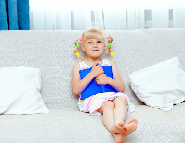 Menina feliz com cabelo loiro está lendo um livro sentado em um sofá na sala de estar de sua casa. ensino à distância.