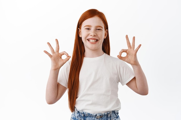 Menina feliz com cabelo ruivo mostra sinais de ok e sorri satisfeita. criança ruiva satisfeita diga sim, gosta e aprova, elogia coisa boa, fundo branco