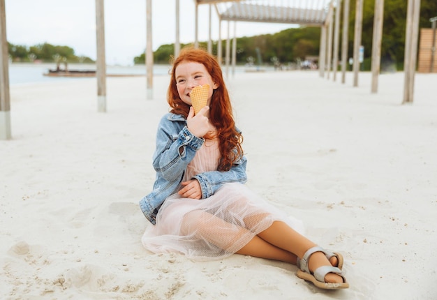 Menina feliz com cabelo vermelho come sorvete na praia nas férias de verão