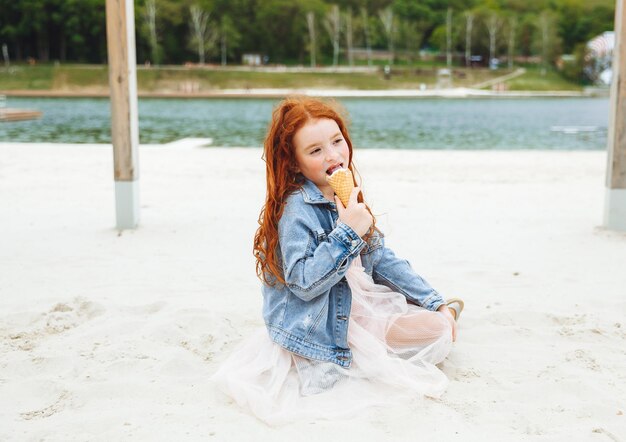 Menina feliz com cabelo vermelho come sorvete na praia nas férias de verão