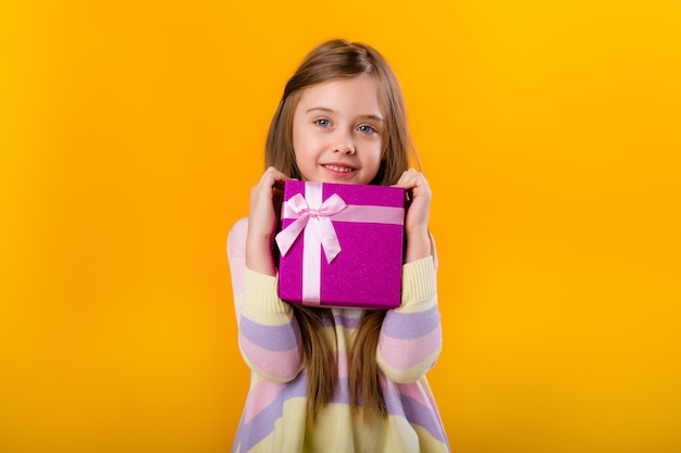 Menina feliz com cabelos longos, segurando uma caixa de presente rosa em um espaço amarelo isolado