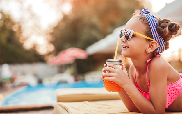 Menina feliz com suco de laranja relaxante à beira da piscina em dia ensolarado de verão