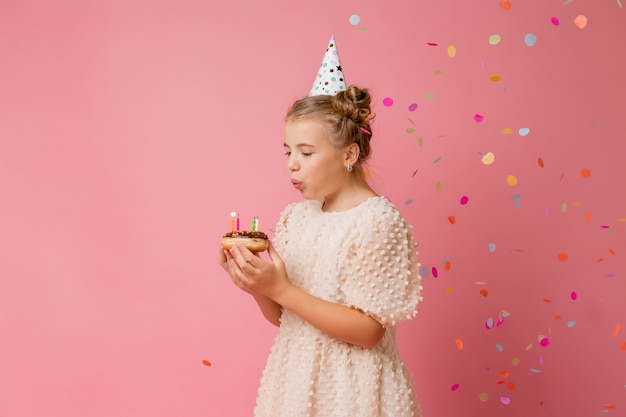 Menina feliz com um boné de aniversário faz um pedido e apaga velas em um bolo.