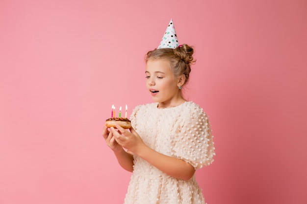 Menina feliz com um boné de aniversário faz um pedido e apaga velas em um bolo.