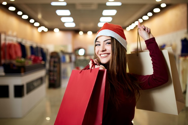 Menina feliz comprando presentes no shopping na venda de natal Conceito de ideia de compras de férias de ano novo Mulher bonita caucasiana sorridente com papel colorido apresenta sacos usando chapéu de natal na loja ou loja