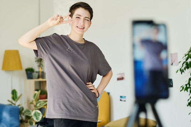 Menina feliz de camiseta mostrando um gesto de paz e olhando para a câmera do smartphone