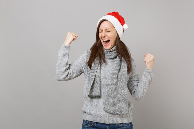 Menina feliz do Papai Noel no chapéu do Natal do cachecol da camisola com os olhos fechados gritando, fazendo o gesto do vencedor isolado no fundo cinza. Conceito de festa de feriado de celebração de ano novo feliz 2019. Simule o espaço da cópia.