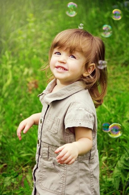 Menina feliz e alegre em um dia ensolarado de verão no contexto da natureza e bolhas de sabão