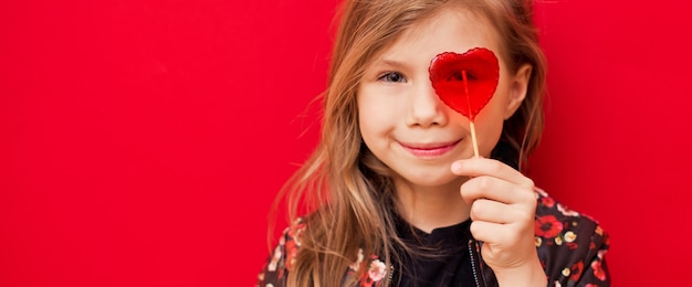 Menina feliz e animada cobriu os olhos com um pirulito em forma de coração brilhante sobre fundo vermelho
