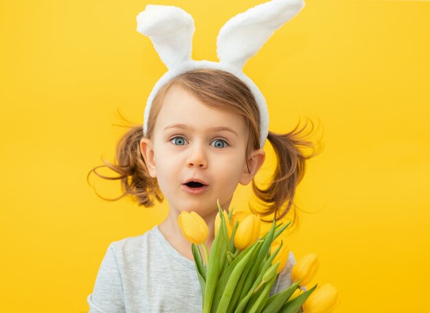 Menina feliz e engraçada com dois rabos de cavalo usando uma tiara com orelhas de coelho, segurando um buquê de tulipas amarelas e a boca aberta