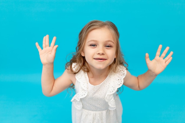 Menina feliz e fofa em um vestido de algodão branco sobre fundo azul no estúdio levantou as mãos e brincou, um lugar para texto