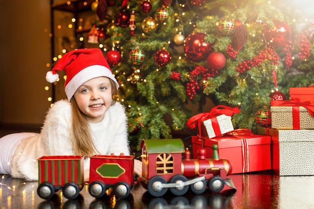 Menina feliz e sorridente com um chapéu de Papai Noel perto de uma caixa de presente mágica e um trem de brinquedo no fundo da árvore de Natal. Feliz Natal