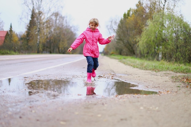 Menina feliz em botas de borracha de jaqueta impermeável rosa alegremente salta através de poças na estrada de rua em tempo chuvoso Primavera outono Diversão infantil ao ar livre depois da chuva Recreação ao ar livre