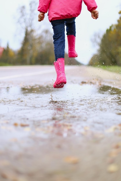 Menina feliz em botas de borracha de jaqueta impermeável rosa alegremente salta através de poças na estrada de rua em tempo chuvoso Primavera outono Diversão infantil ao ar livre depois da chuva Recreação ao ar livre