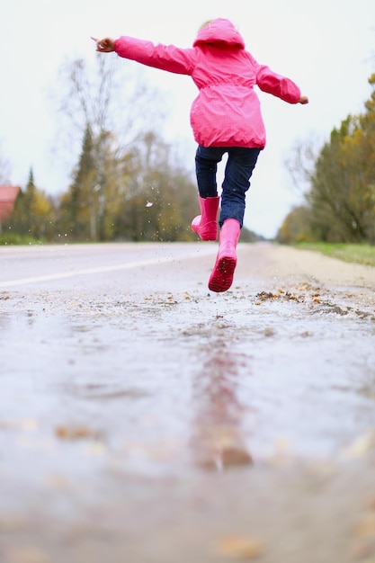 Menina feliz em botas de borracha de jaqueta impermeável rosa alegremente salta através de poças na estrada de rua em tempo chuvoso Primavera outono Diversão infantil ao ar livre depois da chuva Recreação ao ar livre