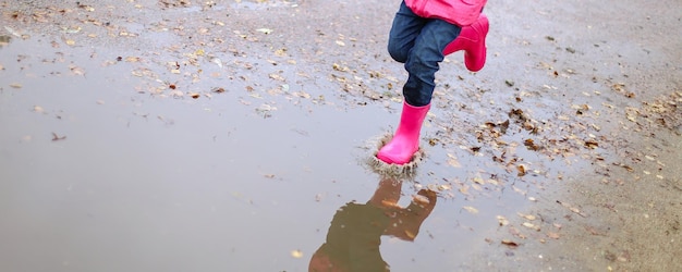 Menina feliz em botas de borracha de jaqueta impermeável rosa alegremente salta através de poças na estrada de rua em tempo chuvoso Primavera outono Diversão infantil ao ar livre depois da chuva Recreação ao ar livre