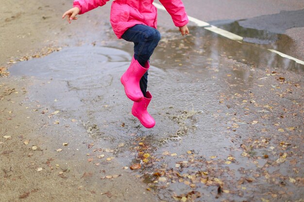 Menina feliz em botas de borracha de jaqueta impermeável rosa alegremente salta através de poças na estrada de rua em tempo chuvoso Primavera outono Diversão infantil ao ar livre depois da chuva Recreação ao ar livre