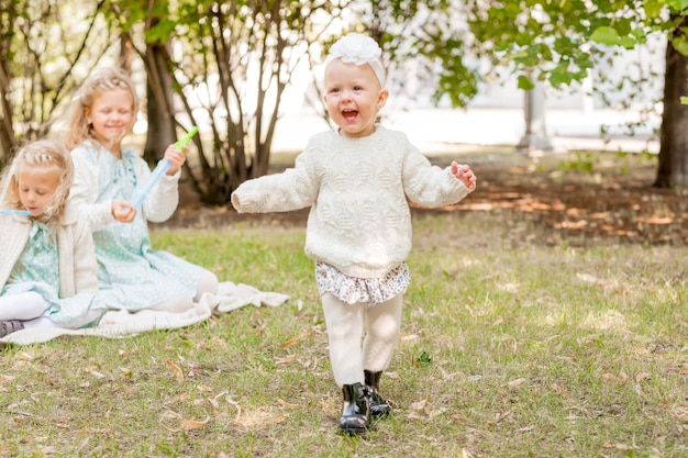 Foto menina feliz em calças brancas e bandas na cabeça em um piquenique com suas irmãs o bebê está aprendendo a andar