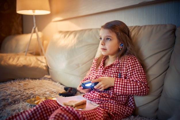 Menina feliz em pajams sentada no sofá, comendo pizza e jogando videogame com joystick em casa