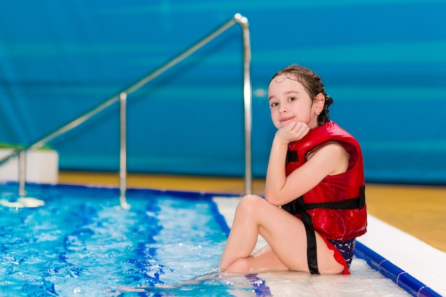 Menina feliz em um colete vermelho, sentado nos degraus da piscina no parque aquático