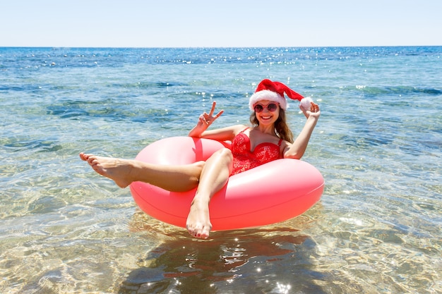 Menina feliz em um donut polvilhado e chapéu de Natal flutuando no mar, sorrindo com óculos de sol para o verão