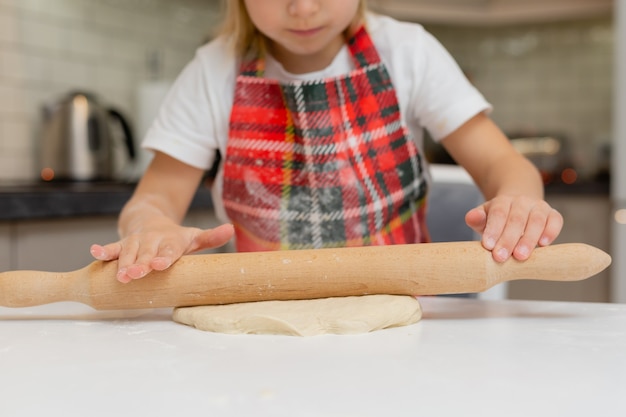 Menina feliz em uma fantasia de chef desenrola a massa com um rolo de massa na cozinha