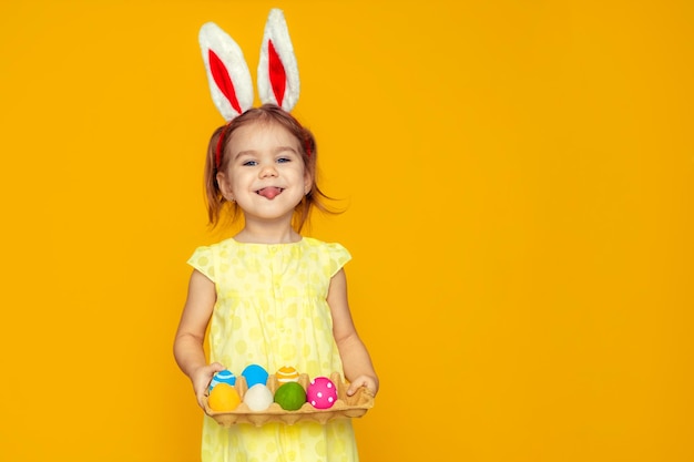 Menina feliz engraçada com ovos de páscoa e orelhas de coelho em fundo amarelo