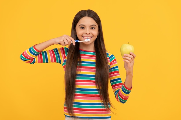 Menina feliz escova os dentes com escova de dentes segurando fundo amarelo maçã, dentes saudáveis.