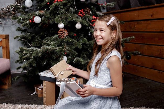 Menina feliz olha para os presentes de Natal perto da árvore de Natal atrás.