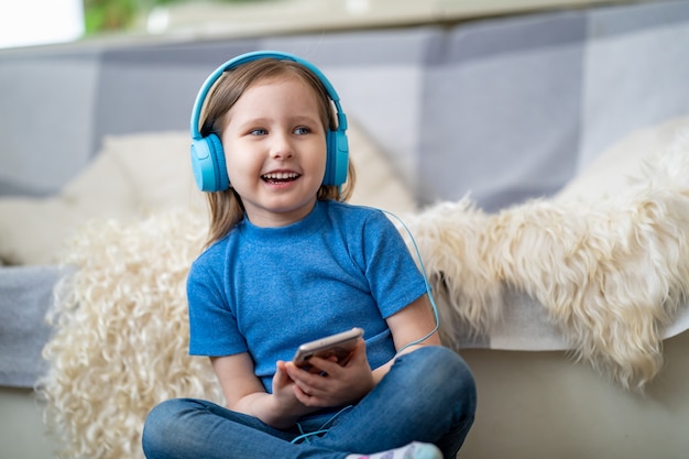 Menina feliz ouvindo música em fones de ouvido azuis, em casa.