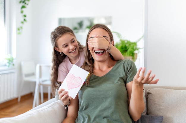 Menina feliz parabenizando a mãe sorridente e dando cartão com coração vermelho durante a celebração do feriado em casa cobrindo os olhos e surpreendendo-a