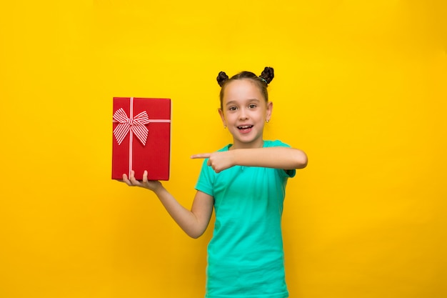 Menina feliz que está isolada sobre o fundo amarelo que guarda a caixa de presente vermelha.