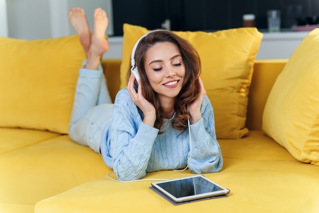 Menina feliz satisfeita usando tablet enquanto estava deitado no confortável sofá amarelo e apreciando músicas agradáveis com fones de ouvido.