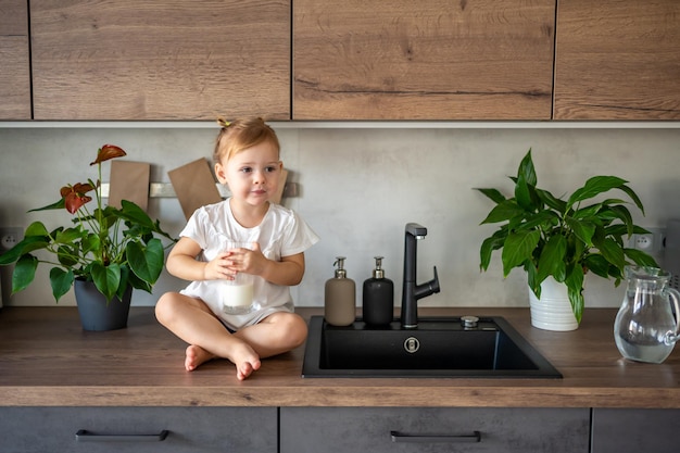 Menina feliz sentada à mesa na cozinha e bebendo leite