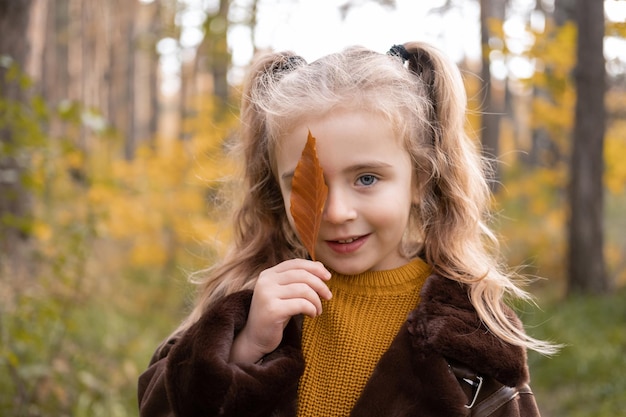 Menina feliz sorrindo, cobrindo o rosto com licença de outono na floresta de outono