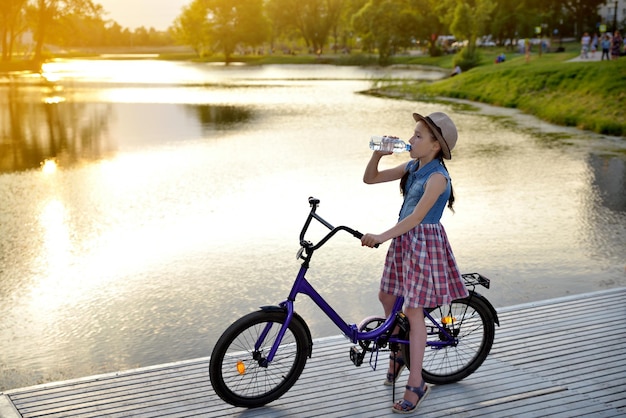 Menina fica com uma bicicleta na margem do rio nos raios solares e bebe água de uma garrafa