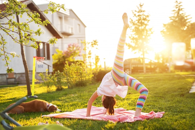 Menina flexível realizando ginástica em um cobertor na grama, enquanto seu cachorro está olhando para ela.