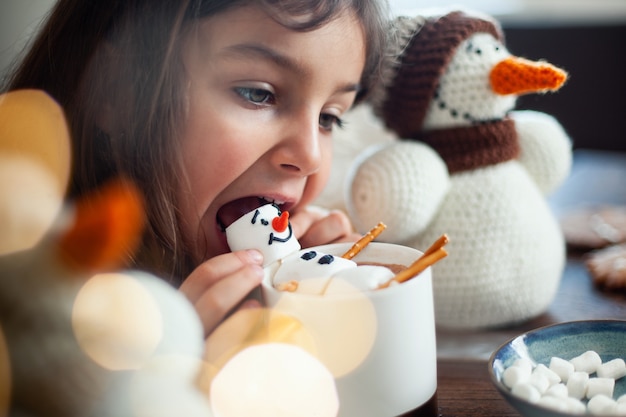 Menina fofa comendo um boneco de neve marmmello decorado com cobertura e bebendo chocolate