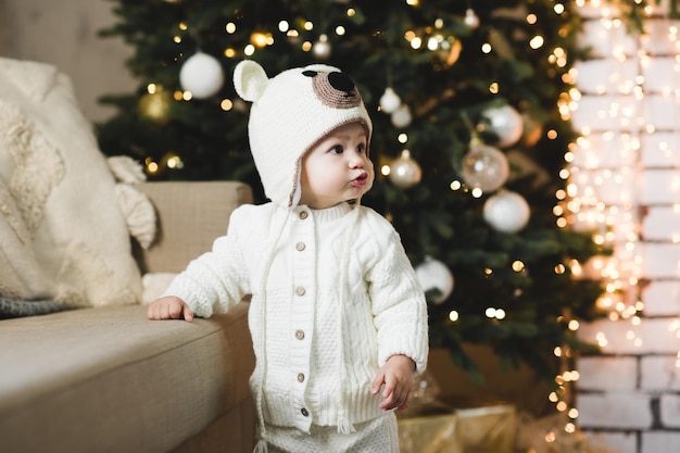 Menina fofa vestindo terno de malha e chapéu posando sobre a árvore de Natal