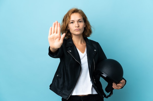 Menina georgiana segurando um capacete de motociclista isolado em um fundo azul, fazendo um gesto de pare