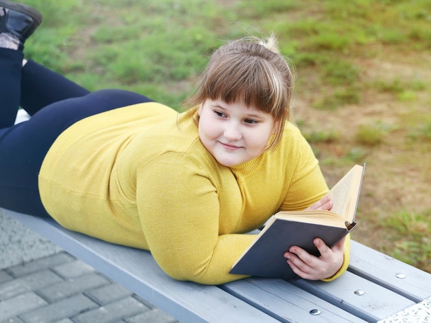 Menina gorda e sorridente, deitada no banco do parque, segura o livro nas mãos e desvia o olhar