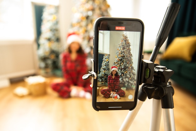 Foto menina gravando seu videoblog de natal por smartphone no tripé. ela está vestindo pijama vermelho e chapéu de papai noel.