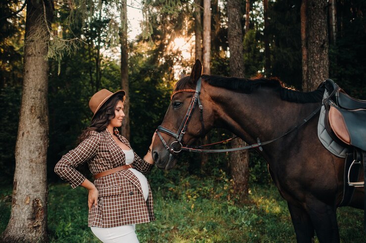 Foto menina grávida com uma grande barriga em um chapéu ao lado de cavalos na floresta na natureza. uma mulher grávida elegante em um vestido branco e jaqueta marrom com cavalos.