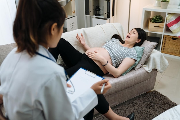 Menina grávida preocupada deitada no sofá ao lado do conselheiro feminino asiático durante a terapia na sala de estar em casa. médico psiquiatra examinando a paciente da maternidade. dando conselhos e sugestões.