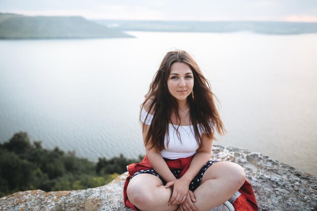 Menina hippie com cabelo ventoso sentado e meditando no topo da montanha rochosa com bela vista do rio Momento calmo atmosférico Viagens e meditação Copiar espaço