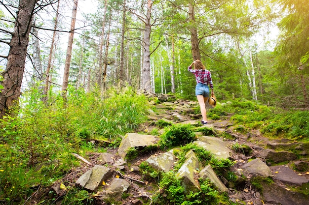 Menina hippie nas montanhas. Mulher elegante com chapéu de palha e camisa quadriculada na floresta. Conceito de Wanderlust. Viajar e fazer caminhadas no verão. madeira misteriosa. Natureza bela.